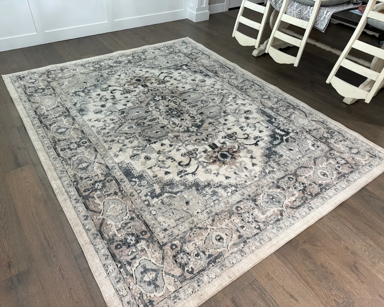 Recently professionally cleaned large patterned area rug in soft gray, cream, and beige tones, looking fresh and restored on a dark wood floor in a bright dining area.